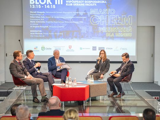 Grupa osob na scenie. Kobieta przemawia przez mikrofon, a pozostali słuchają tego co mówi.