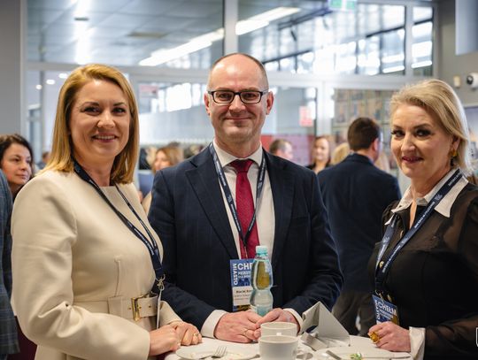 Dwie,kobiety i mężczyzna ubrani w eleganckie stroje pozują do zdjęcia. W tle grupa innych osób.