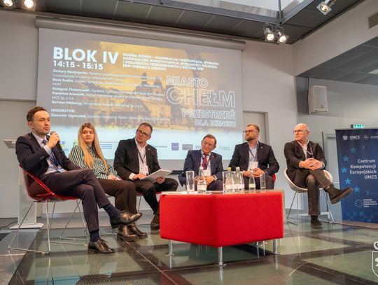 Grupa osób siedzi na scenie. Mężczyzna mówi do mikrofonu, a pozostali go słuchają.