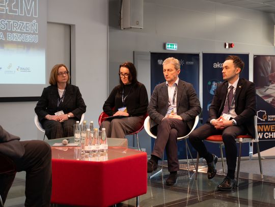 Dwóch mężczyzn i dwie kobiety siedzą na krzesłach na scenie. W tle ekran oraz ścianki promocyjne.